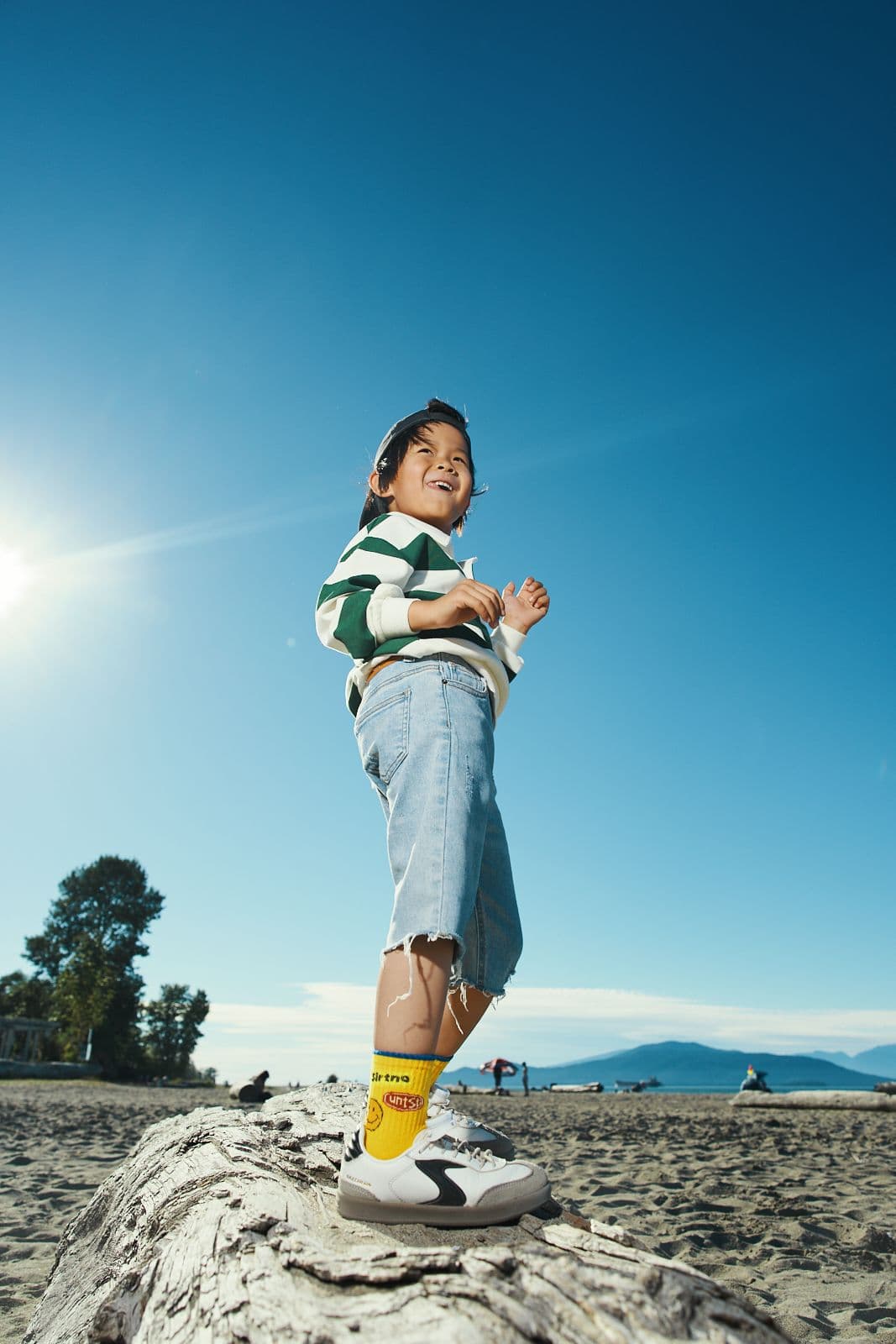 Kids commercial campaign photo of a boy in a striped rugby shirt standing on beach driftwood in backlit sunshine in Vancouver by Una Zhu