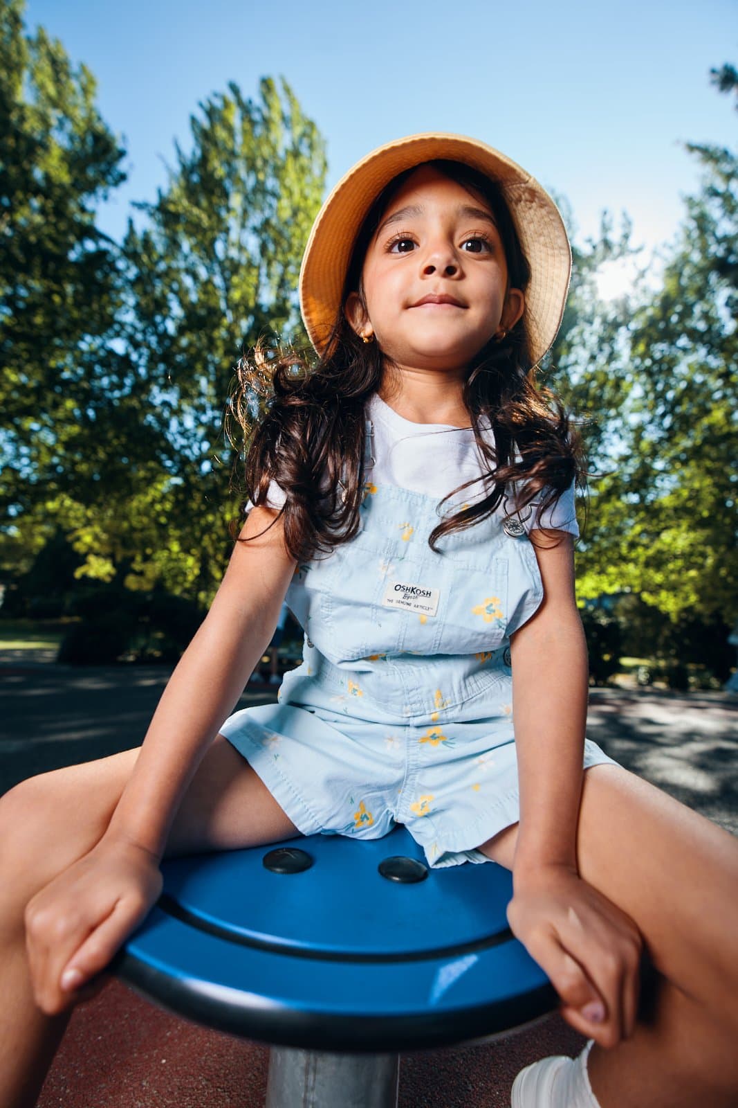 Kids apparel campaign portrait of a girl in a straw sun hat on a playground spinner in Vancouver by Una Zhu